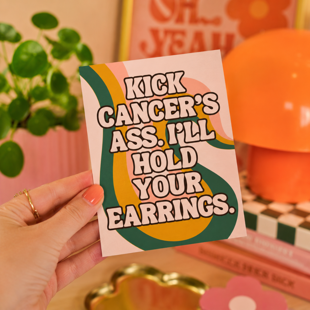 A hand holds a retro-style greeting card with the text 'KICK CANCER'S ASS. I'LL HOLD YOUR EARRINGS.